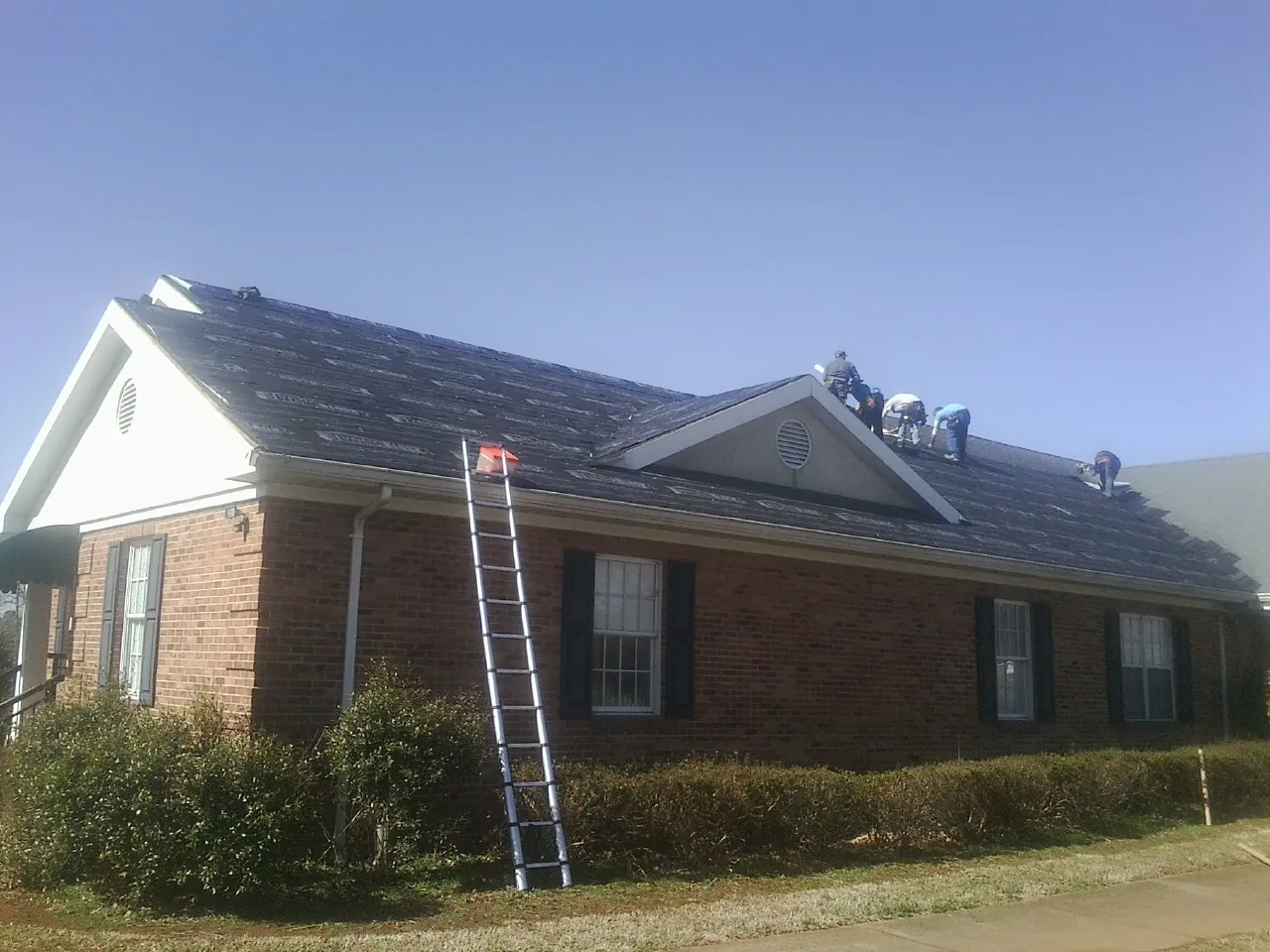 Crew tearing shingles off a brick ranch roof with a ladder set — Emergency Roof Repair work by our crew in Challis