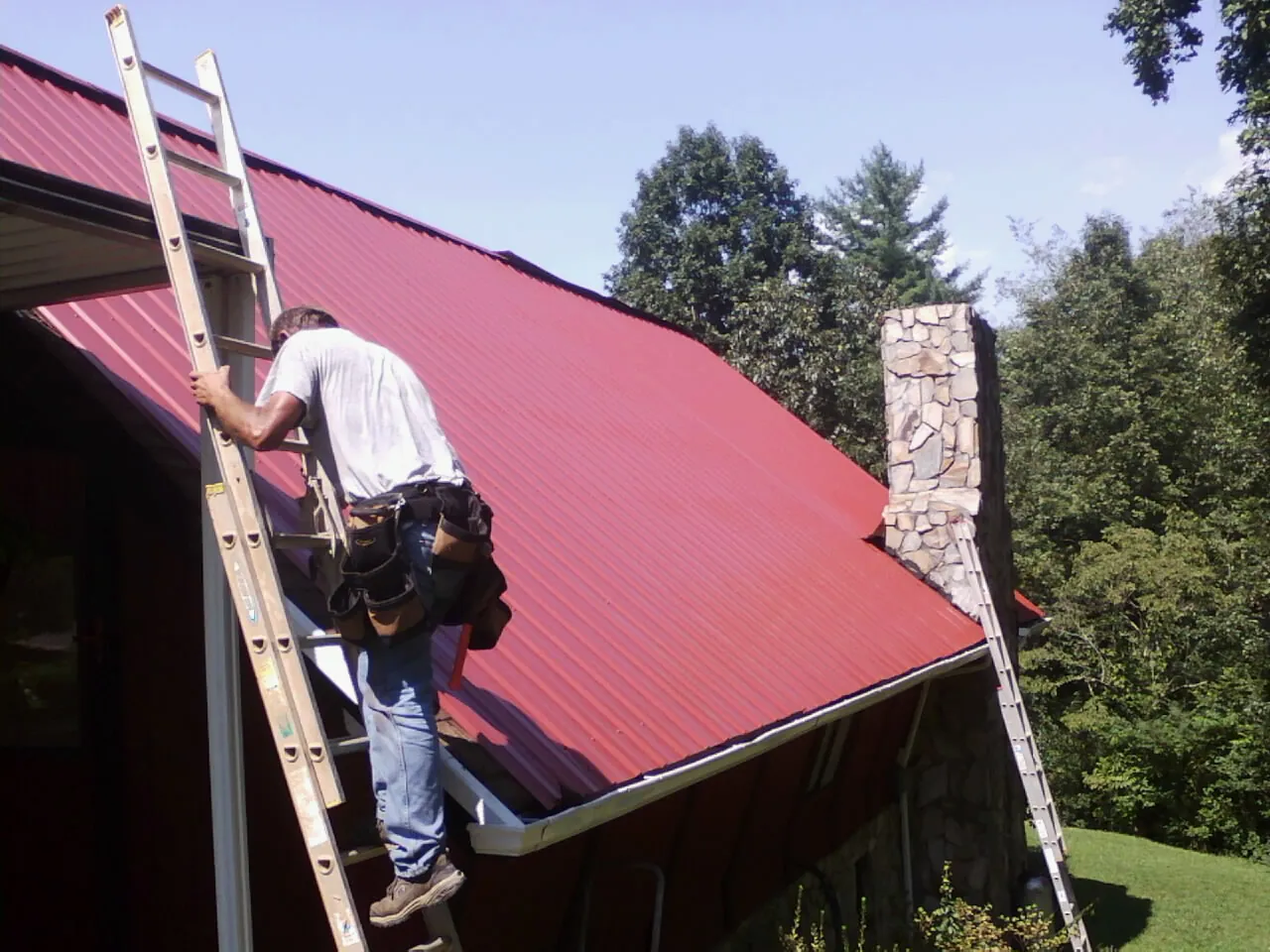 Roofer on a ladder beside a red metal roof and stone chimney — Soffit & Fascia Repair by Roofing Brothers in Challis